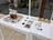 An arrangement of artistic objects on white tables, featuring a central wooden block with a delicate wire sculpture surrounded by broken ceramic pieces. There are several tile samples in various shades, alongside scattered ceramic fragments. The setting appears to be a modern indoor space, with large glass windows and potted plants visible in the background.