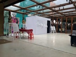 An interior space featuring a modern exhibition setup with a glass ceiling. There are posters and informational boards displayed on stands, and a small table covered with a red cloth is surrounded by red stools. The bright, open setting is accented with plants and decorative elements on the walls.