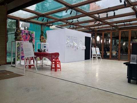 An interior space featuring a modern exhibition setup with a glass ceiling. There are posters and informational boards displayed on stands, and a small table covered with a red cloth is surrounded by red stools. The bright, open setting is accented with plants and decorative elements on the walls.