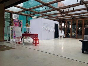An interior space featuring a modern exhibition setup with a glass ceiling. There are posters and informational boards displayed on stands, and a small table covered with a red cloth is surrounded by red stools. The bright, open setting is accented with plants and decorative elements on the walls.