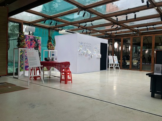 An interior space featuring a modern exhibition setup with a glass ceiling. There are posters and informational boards displayed on stands, and a small table covered with a red cloth is surrounded by red stools. The bright, open setting is accented with plants and decorative elements on the walls.