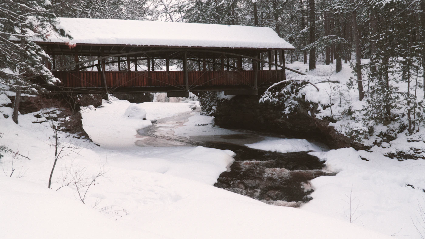 A historic red covered bridge spanning a snow-covered river in winter, surrounded by bare trees and white forest in Ontario