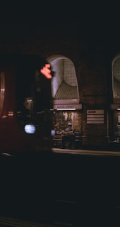 A dimly lit underground train station platform with a person sitting on a bench, reading a newspaper. The station has arched windows and brick walls, with the name 'Baker Street' visible on a sign. A train is passing by in the foreground, slightly blurred due to motion.