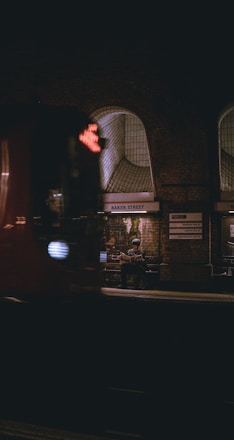 A dimly lit underground train station platform with a person sitting on a bench, reading a newspaper. The station has arched windows and brick walls, with the name 'Baker Street' visible on a sign. A train is passing by in the foreground, slightly blurred due to motion.