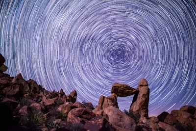 A breathtaking night sky with circular star trails creating a mesmerizing pattern. Below, a rugged landscape features prominently with large, weathered rocks forming a natural arch. The scene exudes a sense of wonder and natural beauty.