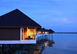 Waterfront bungalows with thatched roofs are built on stilts over a calm body of water. Warm interior lights are visible through the windows, creating a cozy ambiance against the backdrop of a serene, blue sky at dusk.