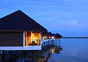 Waterfront bungalows with thatched roofs are built on stilts over a calm body of water. Warm interior lights are visible through the windows, creating a cozy ambiance against the backdrop of a serene, blue sky at dusk.