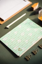A flat lay of stationery items arranged neatly on a textured green surface, featuring a patterned notebook with a geometric design in pastel colors, a white pen, three gold binder clips, a wooden clipboard, and geometric blocks partially visible.