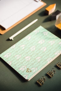A flat lay of stationery items arranged neatly on a textured green surface, featuring a patterned notebook with a geometric design in pastel colors, a white pen, three gold binder clips, a wooden clipboard, and geometric blocks partially visible.