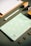 A flat lay of stationery items arranged neatly on a textured green surface, featuring a patterned notebook with a geometric design in pastel colors, a white pen, three gold binder clips, a wooden clipboard, and geometric blocks partially visible.