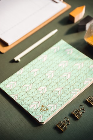 A flat lay of stationery items arranged neatly on a textured green surface, featuring a patterned notebook with a geometric design in pastel colors, a white pen, three gold binder clips, a wooden clipboard, and geometric blocks partially visible.