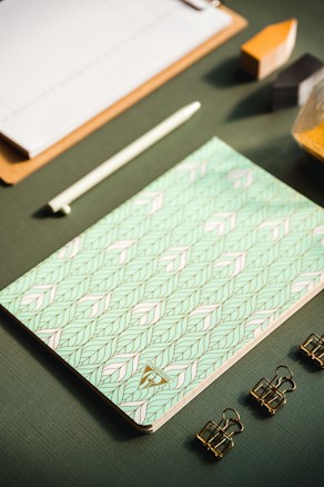 A flat lay of stationery items arranged neatly on a textured green surface, featuring a patterned notebook with a geometric design in pastel colors, a white pen, three gold binder clips, a wooden clipboard, and geometric blocks partially visible.