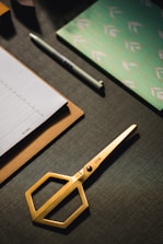A set of neatly arranged stationery items is on a dark textured surface. There is a gold pair of scissors with a modern design, a stack of paper on a clipboard, a pen, and a decorative notebook with a green and white pattern.
