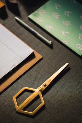 A set of neatly arranged stationery items is on a dark textured surface. There is a gold pair of scissors with a modern design, a stack of paper on a clipboard, a pen, and a decorative notebook with a green and white pattern.