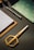 A set of neatly arranged stationery items is on a dark textured surface. There is a gold pair of scissors with a modern design, a stack of paper on a clipboard, a pen, and a decorative notebook with a green and white pattern.