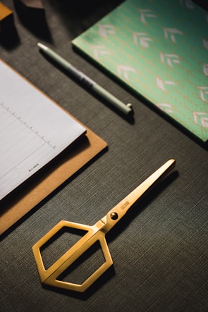 A set of neatly arranged stationery items is on a dark textured surface. There is a gold pair of scissors with a modern design, a stack of paper on a clipboard, a pen, and a decorative notebook with a green and white pattern.