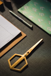 A set of neatly arranged stationery items is on a dark textured surface. There is a gold pair of scissors with a modern design, a stack of paper on a clipboard, a pen, and a decorative notebook with a green and white pattern.