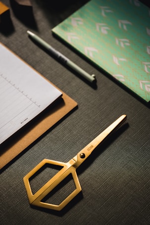 A set of neatly arranged stationery items is on a dark textured surface. There is a gold pair of scissors with a modern design, a stack of paper on a clipboard, a pen, and a decorative notebook with a green and white pattern.