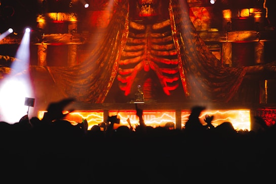 A concert scene with dramatic stage decorations, including large skeletal structures and vivid lighting effects. The stage is illuminated with fiery orange and red colors, creating an intense atmosphere, while a crowd of attendees watches, silhouetted against the light.