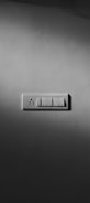 A grayscale photograph of a light switch panel mounted on a plain wall. The panel contains a combination of three switches and one electrical socket. Shadows cast around the panel give it a dramatic look.