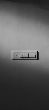 A grayscale photograph of a light switch panel mounted on a plain wall. The panel contains a combination of three switches and one electrical socket. Shadows cast around the panel give it a dramatic look.