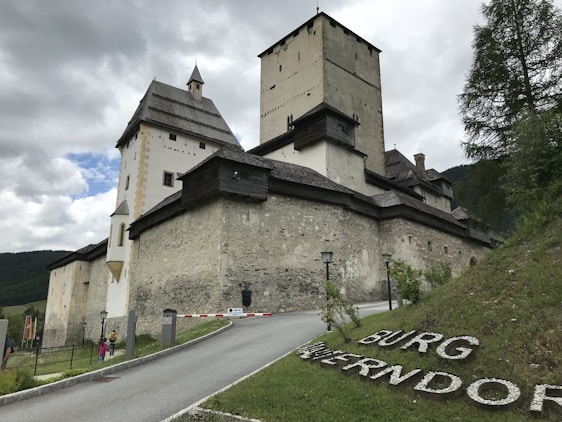 A large, historic stone castle with multiple towers and a steep roof, set against a cloudy sky. The castle is surrounded by lush greenery and a paved path leading uphill. There are two people walking near the entrance, and letters spelling 'BURG WERFEN' are displayed on the grassy slope.