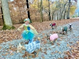 A young girl dressed in a colorful costume, possibly resembling a fairy, dances playfully on a gravel path in a forest with autumn leaves scattered around. Accompanying her are several small goats, each wearing cute sweaters in different patterns and colors.