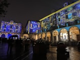 A nighttime scene of a square with buildings illuminated by colorful, celestial-themed projections. Stars, planets, and other astronomical elements are displayed on the building facades, creating a vibrant and artistic atmosphere. People with umbrellas can be seen walking on the wet cobblestone, suggesting recent rain. Festive lights add to the ambiance, highlighting the arches and windows.