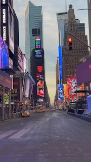 A view of a city street lined with tall buildings adorned with bright, colorful digital advertisements and billboards. The street is relatively empty, with a few yellow taxis and cars visible on the road. The sky above the cityscape is partly cloudy, and a festive 'Happy New Year 2023' sign is prominently displayed on one of the buildings.