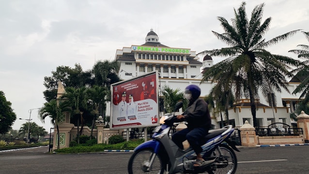 A motorcyclist rides past an official building with a large billboard in the foreground. The building has a dome and multiple stories, surrounded by palm trees and greenery. The billboard displays information related to an upcoming election.