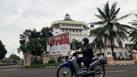 A motorcyclist rides past an official building with a large billboard in the foreground. The building has a dome and multiple stories, surrounded by palm trees and greenery. The billboard displays information related to an upcoming election.