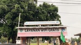 A structure with a corrugated metal roof is situated outdoors, featuring a prominent sign that reads 'Layanan Wifi Gratis' in large letters. Additional text in smaller letters indicates the involvement of a governmental department in the local area, Dinas Komunikasi Informatika dan Statistik Kabupaten Ponorogo. The structure is surrounded by trees and other outdoor elements.