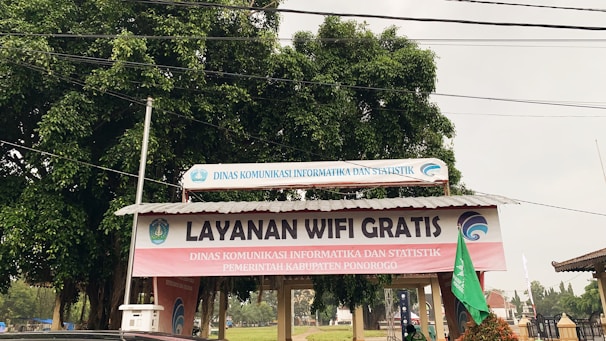 A structure with a corrugated metal roof is situated outdoors, featuring a prominent sign that reads 'Layanan Wifi Gratis' in large letters. Additional text in smaller letters indicates the involvement of a governmental department in the local area, Dinas Komunikasi Informatika dan Statistik Kabupaten Ponorogo. The structure is surrounded by trees and other outdoor elements.
