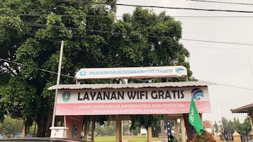 A structure with a corrugated metal roof is situated outdoors, featuring a prominent sign that reads 'Layanan Wifi Gratis' in large letters. Additional text in smaller letters indicates the involvement of a governmental department in the local area, Dinas Komunikasi Informatika dan Statistik Kabupaten Ponorogo. The structure is surrounded by trees and other outdoor elements.
