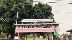 A structure with a corrugated metal roof is situated outdoors, featuring a prominent sign that reads 'Layanan Wifi Gratis' in large letters. Additional text in smaller letters indicates the involvement of a governmental department in the local area, Dinas Komunikasi Informatika dan Statistik Kabupaten Ponorogo. The structure is surrounded by trees and other outdoor elements.