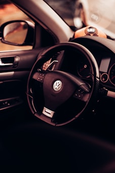 A close-up view of a car interior featuring a Volkswagen steering wheel. The dashboard and part of the driver's side door are visible, with a warm, ambient lighting casting a cozy feel. A golf-themed decorative item is placed on the dashboard.