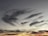 Wispy, feathery clouds stretch across the sky, creating a dynamic and dramatic pattern. The sky transitions from bright orange and yellow near the horizon to deep blues and grays towards the top, suggesting a sunset or sunrise.