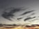 Wispy, feathery clouds stretch across the sky, creating a dynamic and dramatic pattern. The sky transitions from bright orange and yellow near the horizon to deep blues and grays towards the top, suggesting a sunset or sunrise.