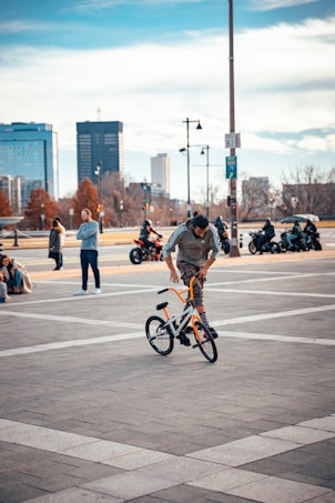A person performs a trick on a BMX bike in an urban setting. The scene includes several people in the background, motorbikes, and a distant city skyline with high-rise buildings. The area is a large, open square with a tiled surface. The sky is partially cloudy, suggesting a cool, pleasant day.