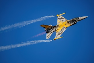 A fighter jet with a striking tiger stripe design in black, gray, and yellow colors flies through a clear blue sky, leaving a trail of white smoke. The jet appears to be ascending at a sharp angle with afterburners firing.