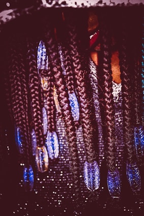 Intricate braids adorned with blue beads hanging down, with a sparkling, textured background. A hint of a person's face partially covered by the braids.