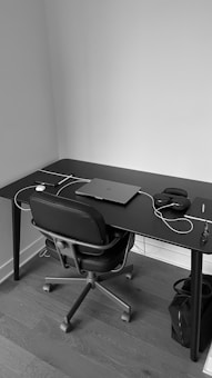 A monochrome image depicts a minimalist workspace with a black desk and an office chair. On the desk, there is a closed laptop, a pair of headphones, a mouse, and some cables. A small waste bin is positioned underneath the right side of the desk. The setting appears organized and tidy, with a simple, modern aesthetic.