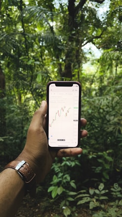 A hand holding a smartphone displaying a stock market chart in the foreground, set against a lush, green forest background. The hand wears a brown leather watch, and the dense foliage creates a natural, serene setting.