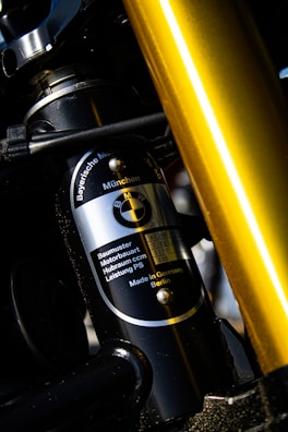 Close-up view of a component detail from a machinery or vehicle, featuring a metallic cylinder with the BMW logo and specifications written on it. The surrounding parts appear to be constructed from metal with visible screws and joints. A prominent golden rod or pipe is also visible in the foreground.