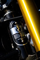 Close-up view of a component detail from a machinery or vehicle, featuring a metallic cylinder with the BMW logo and specifications written on it. The surrounding parts appear to be constructed from metal with visible screws and joints. A prominent golden rod or pipe is also visible in the foreground.