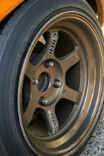 A close-up view of a metallic alloy wheel featuring a five-spoke design. The wheel is mounted on a car with a visible tire surrounding it. The surface of the wheel is slightly worn, indicating use, and branding logos are present on the spokes.