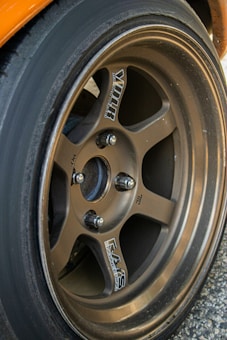 A close-up view of a metallic alloy wheel featuring a five-spoke design. The wheel is mounted on a car with a visible tire surrounding it. The surface of the wheel is slightly worn, indicating use, and branding logos are present on the spokes.