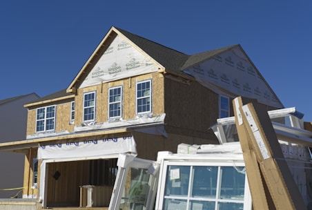 A house under construction with visible framing and insulation wrap. The structure is partially covered with a green guard barrier and has several large windows installed and some stacked in front. Construction materials and debris are scattered in the foreground.