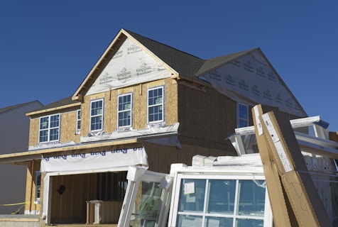A house under construction with visible framing and insulation wrap. The structure is partially covered with a green guard barrier and has several large windows installed and some stacked in front. Construction materials and debris are scattered in the foreground.