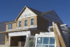A house under construction with visible framing and insulation wrap. The structure is partially covered with a green guard barrier and has several large windows installed and some stacked in front. Construction materials and debris are scattered in the foreground.